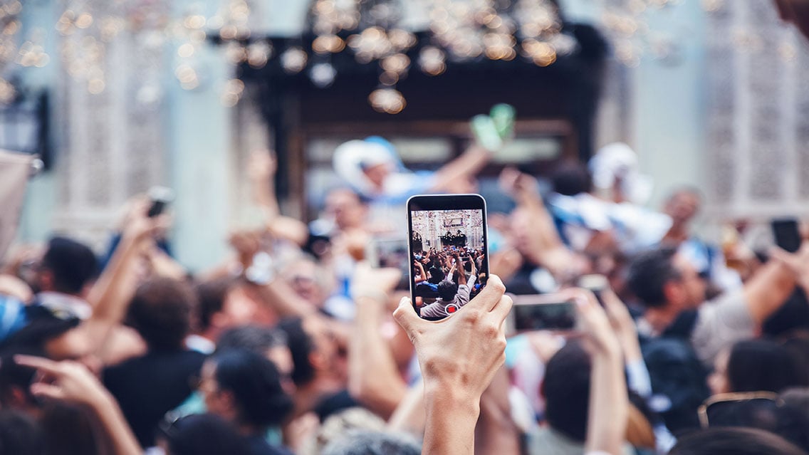 Person at a concert holds phone vertically while recording video for social media in a large crowd